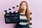 Young beautiful redhead woman holding video film clapboard scared and amazed with open mouth for surprise, disbelief face