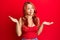 Young beautiful redhead woman wearing casual clothes over red background clueless and confused with open arms, no idea and