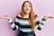 Young beautiful redhead woman wearing striped sweater over pink background clueless and confused with open arms, no idea and