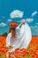 Young beautiful redhead woman in a white shirt with a poppy wreath on her head standing backwards in a poppy field on