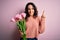 Young beautiful romantic woman with curly hair holding bouquet of pink tulips pointing finger up with successful idea