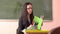 Young beautiful teacher with glasses sits in front of the blackboard and looks into a notebook.