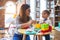 Young beautiful teacher and toddler playing with plastic food and cuttlery toys at kindergarten