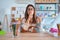 Young beautiful teacher woman wearing sweater and glasses sitting on desk at kindergarten asking to be quiet with finger on lips