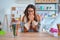 Young beautiful teacher woman wearing sweater and glasses sitting on desk at kindergarten laughing and embarrassed giggle covering