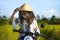 Young beautiful tourist or nomad traveler black afro American woman riding motorbike in tropical field wearing traditional Asian h