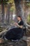 Young, beautiful woman in a black medieval dress with a steel rose in her hands, sitting in the woods on the roots of a tree. Attr