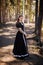 Young, beautiful woman in a black medieval dress walks through the gloomy pine forest.