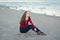 Young beautiful woman with closed eyes, long hair, in black jeans and red shirt, sitting on sand on beach among seagulls birds