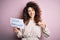 Young beautiful woman with curly hair and piercing holding paper with love yourself message with surprise face pointing finger to