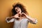 Young beautiful woman with curly hair and piercing wearing striped shirt and glasses smiling in love doing heart symbol shape with