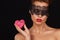 Young beautiful woman with dark lace on eyes bare shoulders and neck, holding cake shape of heart to enjoy the taste and are