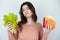 Young beautiful woman holding green salad in one hand and burger with fried potatoes in another looking frustrated being on diet