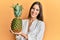 Young beautiful woman holding pineapple looking positive and happy standing and smiling with a confident smile showing teeth