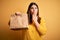 Young beautiful woman holding take away paper bag from delivery over yellow background cover mouth with hand shocked with shame