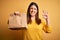 Young beautiful woman holding take away paper bag from delivery over yellow background doing ok sign with fingers, excellent