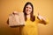 Young beautiful woman holding take away paper bag from delivery over yellow background happy with big smile doing ok sign, thumb