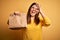 Young beautiful woman holding take away paper bag from delivery over yellow background with happy face smiling doing ok sign with