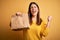 Young beautiful woman holding take away paper bag from delivery over yellow background screaming proud and celebrating victory and