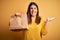 Young beautiful woman holding take away paper bag from delivery over yellow background very happy and excited, winner expression