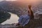 Young beautiful woman in long dress and hat, sits on hill on river background. Evening city sunset