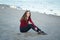 young beautiful woman with long hair, in black jeans and red shirt, sitting on sand on beach among seagulls birds