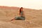 Young beautiful woman with long hair in blue dress sitting elegant and thoughtful on sand at sunrise in sandy desert