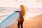 Young beautiful woman with long hair. Surf girl in wetsuit with surfboard on a beach at sunset or sunrise.