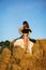 Young beautiful woman looks at the setting sun and holds a straw hat, sits on a large pile of straw bales