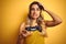 Young beautiful woman playing video game using gamepad over yellow  background stressed with hand on head, shocked with