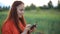 Young beautiful woman in red dress with red hair. watch your photos on your phone and stands in a green wheat field in