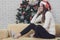 Young beautiful woman in red hat sitting on sofa between christmas trees and listening to music with headphone in front of hugh c