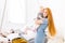 Young beautiful woman with red long hair in a blue dress holds a child on her hands one year blonde near the bed on which lies a m