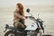 young beautiful woman sitting on her old cafe racer motorcycle in desert at sunset or sunrise