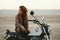 Young beautiful woman sitting on her old cafe racer motorcycle in desert at sunset or sunrise