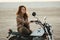 Young beautiful woman sitting on her old cafe racer motorcycle in desert at sunset or sunrise