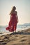 Young beautiful woman walking on beach in red summer dress. Rear view of woman by sea. Fashion, summertime, lifestyle concept