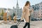 Young beautiful woman walks along city street with travel suitcase and cell phone. Fashionable brunette girl smiling