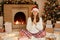Young beautiful woman wearing Santa hat over background of fir tree and fireplace, begging and praying with hands together, with