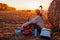 Young bicyclist having rest after a ride in autumn field at sunset. Woman eating by haystack