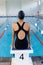 Young biracial female swimmer sitting on starting block at an indoor pool