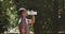 Young black athlete guy taking break in jog workout, drinking water from sport bottle at green summer park, side view