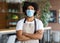 Young Black Barista Wearing Apron And Medical Mask Posing In Cafe Interior