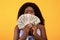 Young black lady covering her face with fan of money, demonstrating shopping budget over orange background