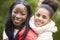 Young black woman piggybacking her pre-teen daughter in the park, both smiling to camera, close up