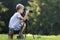 Young blond child boy sitting on tree stump on grassy clearing taking picture with tripod camera