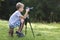 Young blond child boy sitting on tree stump on grassy clearing taking picture with tripod camera