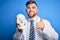 Young blond doctor man with beard and blue eyes wearing coat and glasses holding skull happy with big smile doing ok sign, thumb