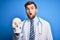 Young blond doctor man with beard and blue eyes wearing coat and stethoscope holding skull scared in shock with a surprise face,