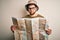 Young blond explorer man with beard and blue eyes wearing glasses and hat holding city map with a confident expression on smart